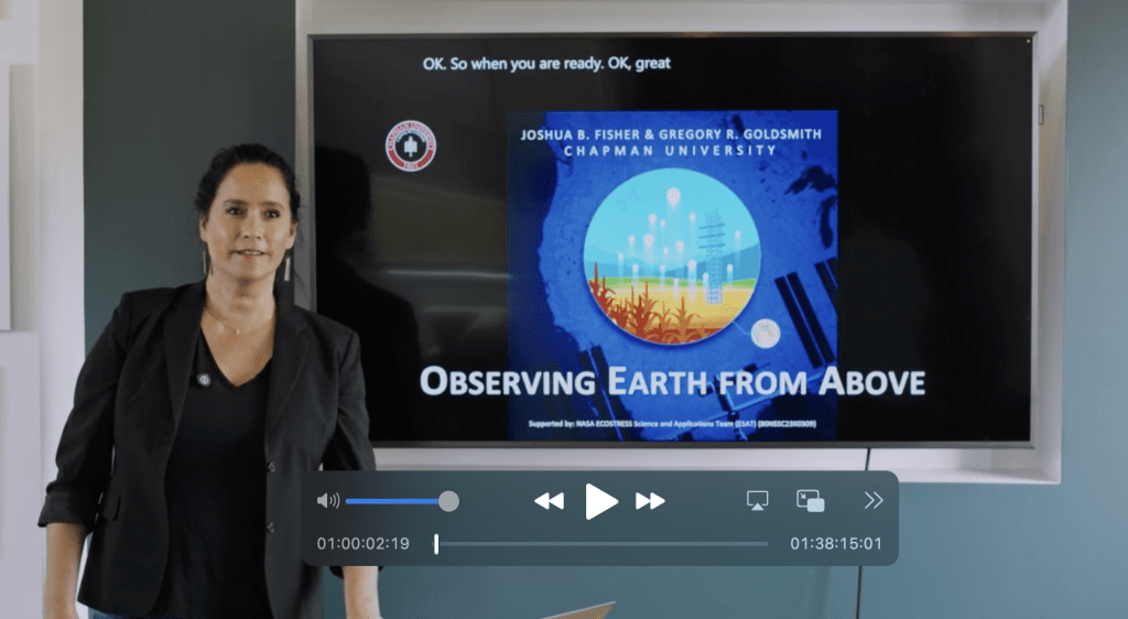 Screenshot of video presentation showing Claudine in a black shirt and black blazer standing in front of a large screen. On the screen is an opening title "Observing Earth from Above," Chapman University logo, and the program sponsoring names Joshua B Fisher and Gregory Goldsmith with captions in the background.